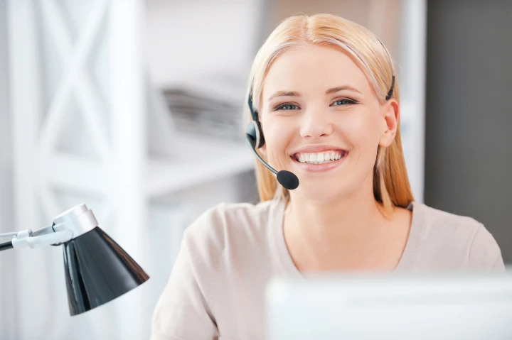 Cosmopolitan Aesthetics Hannover und Braunschweig - Glückliche junge Frau mit Headset, die in die Kamera schaut und lächelt, während sie an ihrem Arbeitsplatz sitzt.
